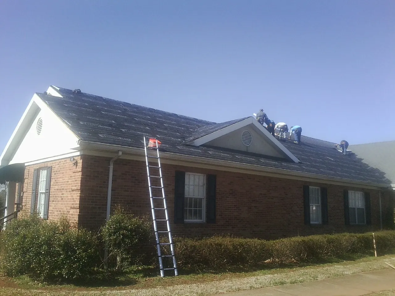 Crew tearing shingles off a brick ranch roof with a ladder set — Metal Roof Repair work by our crew in Smiths Creek