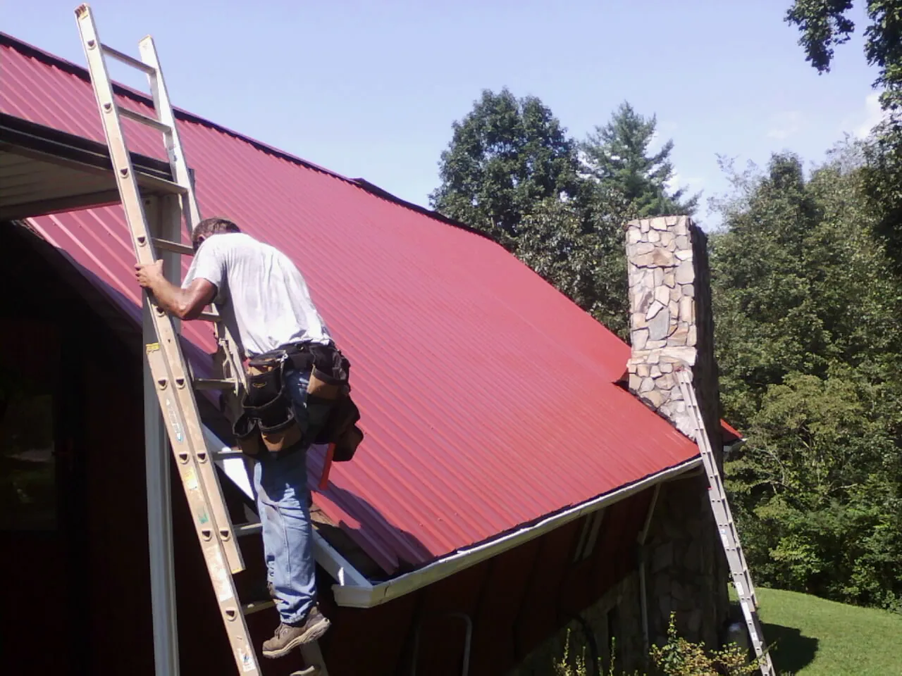 Roofer on a ladder beside a red metal roof and stone chimney — Soffit & Fascia Repair by Roofing Brothers in Smiths Creek